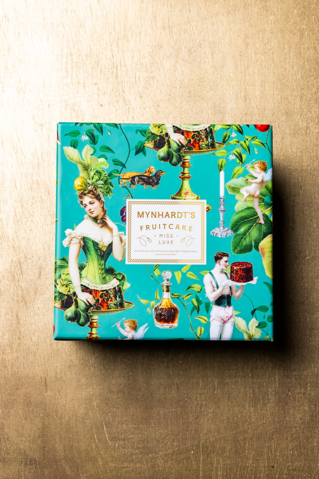 A single Miss Luxe fruitcake box in teal illustrated wrapping, photographed on a brass surface